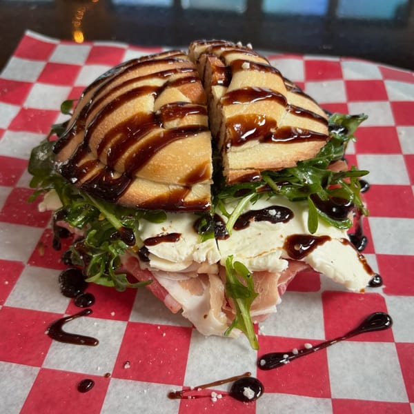 A sliced sandwich with mozzarella, prosciutto, arugula, and balsamic glaze on red and white checkered paper.