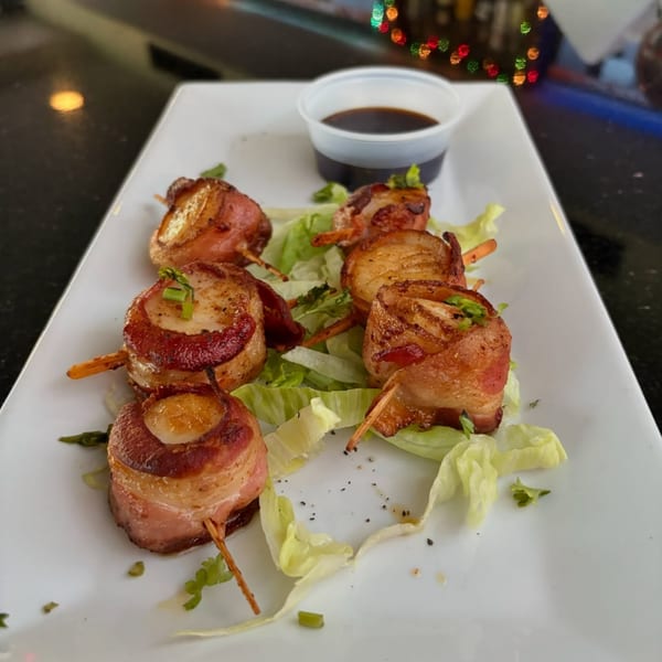 Six bacon-wrapped scallops on skewers with lettuce and a side of dipping sauce on a white plate.
