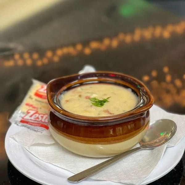 Creamy clam chowder in a brown and tan bowl with crackers and a spoon on a white plate.